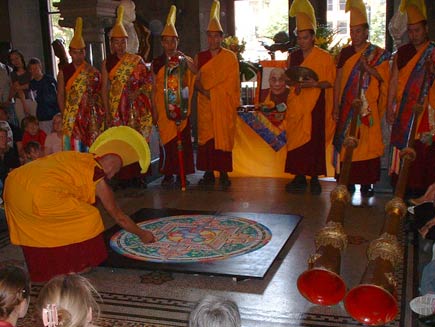 The monks are in full ceremonial dress now, wearing orange robes over their red tunics and orange and yellow crested headdresses. Tall Tibetan horn instruments are also visible.  One monk has begun destroying the mandala by scraping his knuckle in a radial direction through the sand