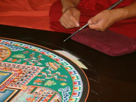 A closer view of the chak-pur rod.  It is a metal tube about the length of the monk's forearm and the thickness of a pen, ridged along its side and tapered towards the end from which the sand is pouring.  The monk is scraping a smaller rod along the chak-pur to make the sand flow smoothly
