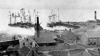 Black and white view over rooftops and industrial chimneys to sailing boats in harbour.