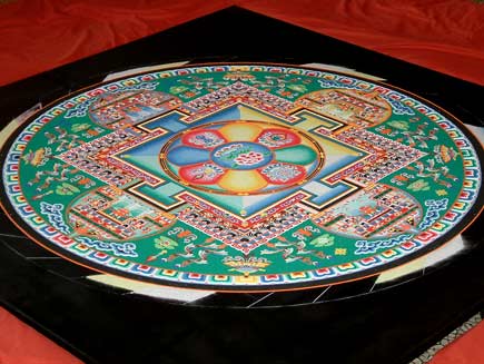 A view of the mandala, now close to completion. The pattern contains squares and circles and is symmetrical in four directions.  The dominant colour is green, with red, blue and yellow sand and many details in black and white.  The middle section represents a building and green areas nearer the edge resemble gardens filled with trees and other designs