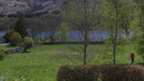 Colour view of an untended garden bounded by stone walls, set next to a narrow loch with tall hills in the background. Aside from a small hedge, some broken-down walls and three tall trees, the garden is given over to grass. To the left, a woman in a red anorak stands on an overgrown path.