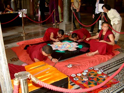 A woman peeks over the shoulder of one of the monks.  They are working in a cordoned-off area inside Manchester Town Hall, attracting one or two spectators