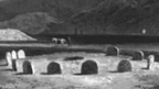 Black and white engraving of Temple Wood stone circle, showing eleven short vertical slabs aranged around a central hollow. To the rear is a taller mound or cairn with high hills behind.