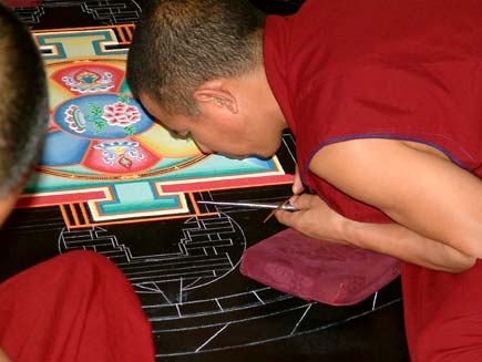 Monks, dressed in red tunics with close-cropped hair, lean over a circular design marked out in chalk on a black tabletop.  Their faces are inches from the surface.  Resting their hands on small cushions, they are pouring coloured sand through small metal tubes into the spaces marked by the chalk outline